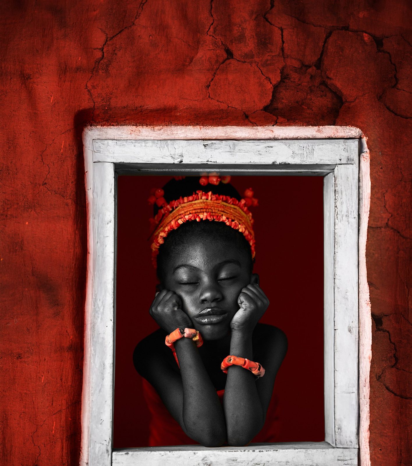 A girl in a coral crown rests her face on her fists inside a white window frame set in a red-earth wall